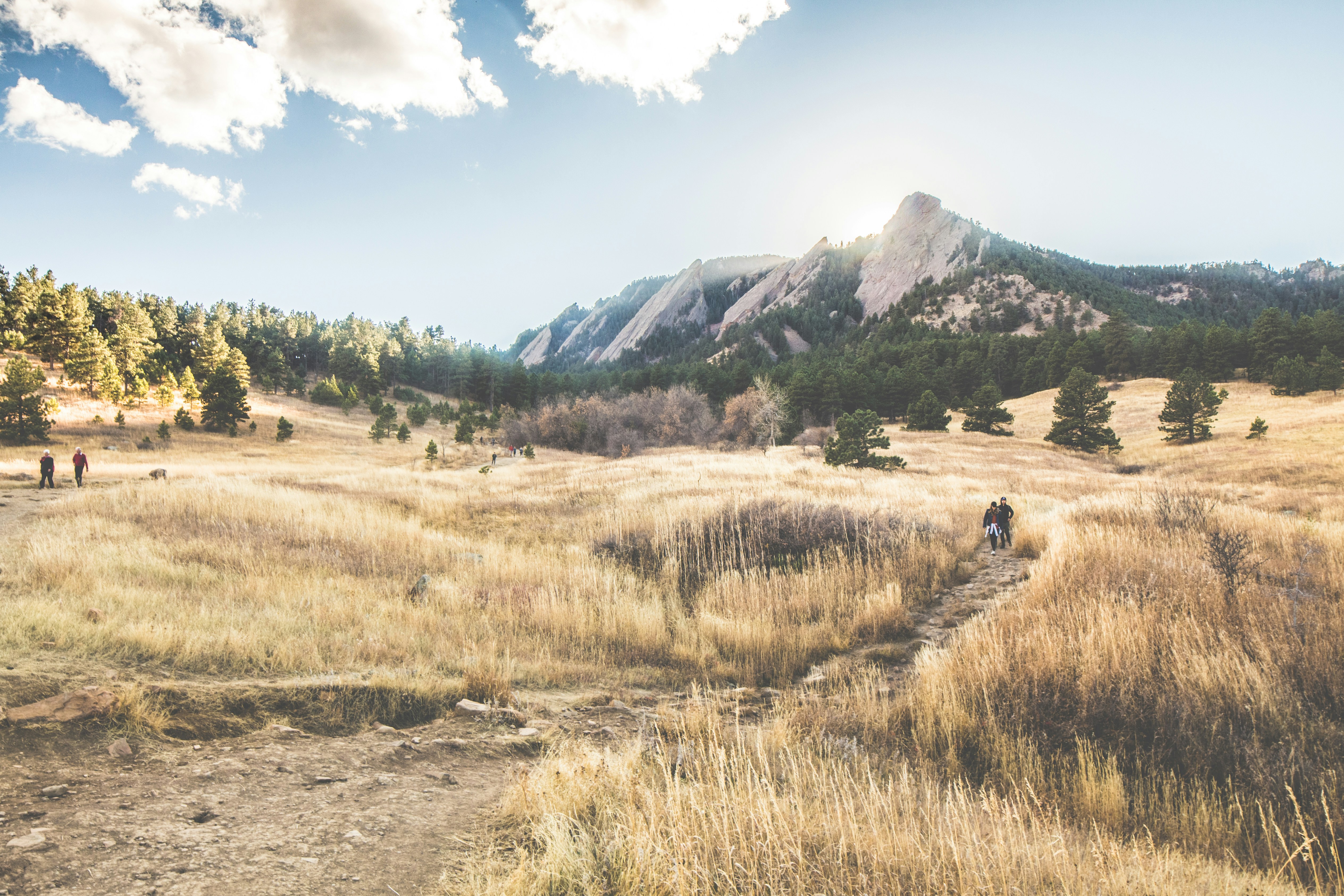 A view of Flagstaff mountain in the distance with light blue skies. The fields of grass are dry and golden at this time of year. There's a dirt trail with a few hikers in the distance.
